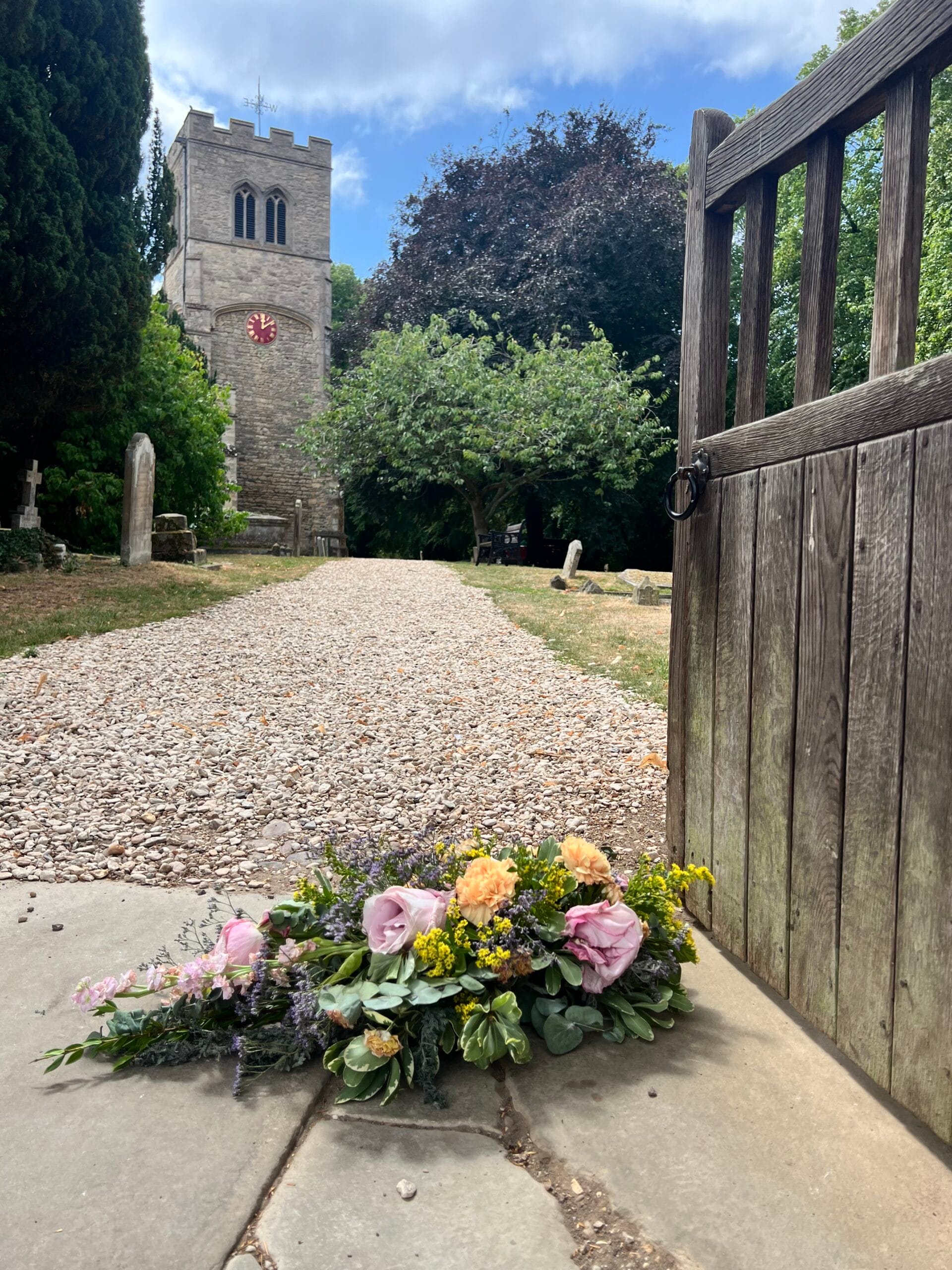 Single ended funeral spray in soft pinks and yellows at a churchyard gate